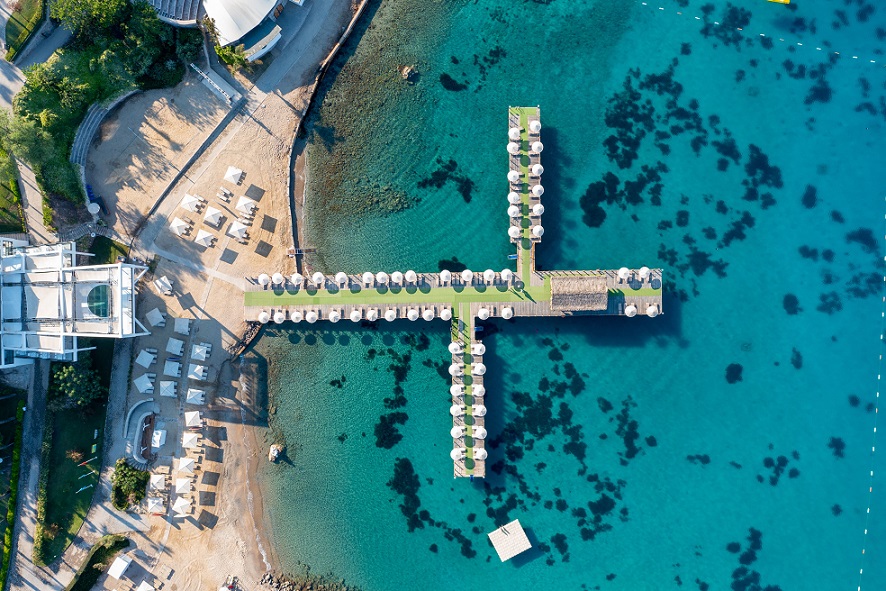 Urlaub Türkei 5 Sterne all inclusive mit Flug -bei REISEBÜRO Wache, Erfurt buchen: Bild aus der Vogelperspektive auf einen Steg mit zwei Seitenarmen, auf dem Sonnenschirme sind, rundherum blaues Meer, davor beigefarbener Strand und ebenfalls Sonnenschirme in der Sonne