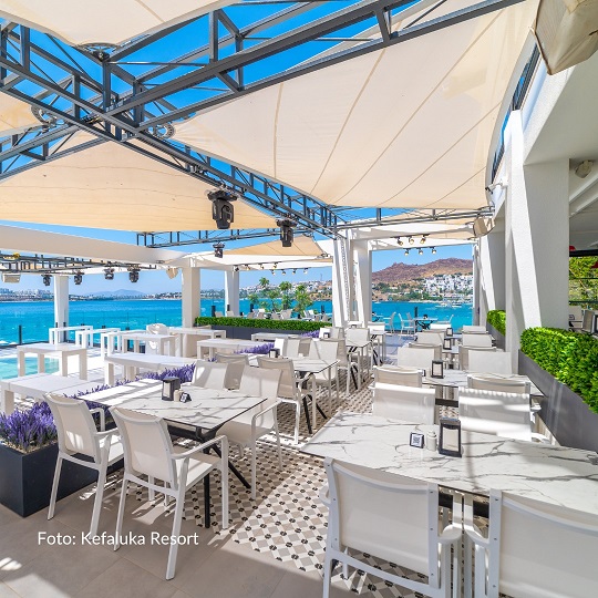 Erfurt REISEBÜRO Wache Türkei Urlaub 5-Sterne-Hotel, im Bild: Blick auf die Terrasse eines Restaurants im 5-Sterne-Hotel, zu sehen: Tische mit weißen Decken, weißen Stühlen, Gläsern und weißem Sonnenschutz über der Terrasse, in der Ferne das Meer