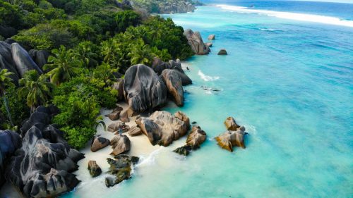 Kreuzfahrt Seychellen buchen Erfurt, REISEBÜRO Wache; Bild von oben: von links Bäume und Vegetation in kräftigem Grün, an dessen Rand große, dunkelgraue Kiesel, etwas Sandstrand, türkisblaues Meer