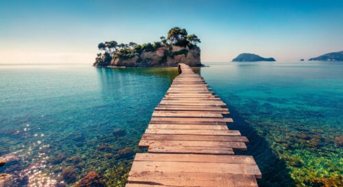 Urlaub Griechenland buchen bei REISEBÜRO Wache Erfurt; im Bild: langer Holzsteg durchs Meer zu einer mit Bäumen bewachsenen Insel unter blauem Himmel auf der Insel Zakynthos